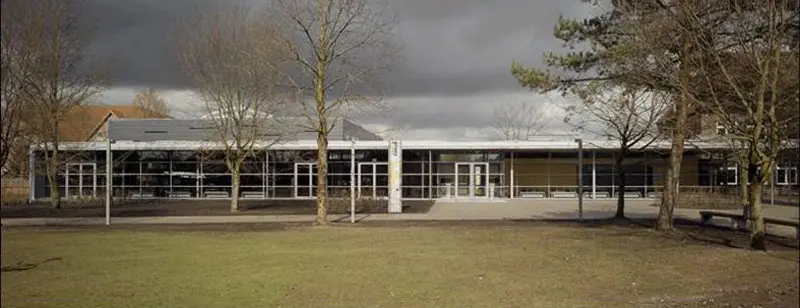 Moderne Schulgebäude der AMG Friesoythe mit Glasfront, umgeben von kahlen Bäumen und einer großen Rasenfläche unter bewölktem Himmel