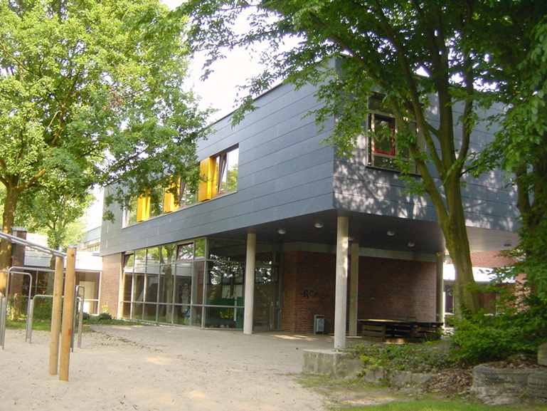 Moderner Schulbau mit großen Fenstern, umgeben von hohen Bäumen und Sandfläche, unter blauem Himmel