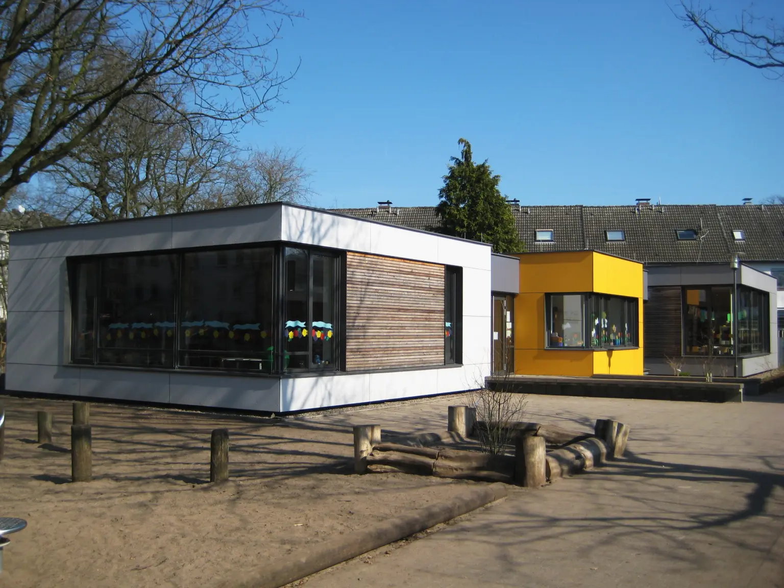 Moderne, farbenfrohe Kindertagesstätte mit großen Fenstern und Spielplatz, umgeben von Bäumen im Sonnenschein
